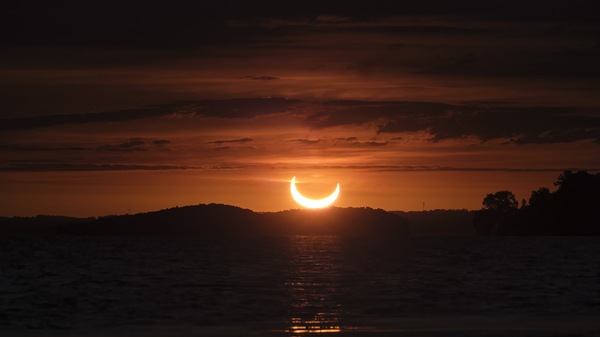 Between layers of clouds, a partial solar eclipse is seen at sunrise over Rice Lake, Ontario, as the sun, partially covered by the moon, rises in a beautiful orange sky above the trees and water.